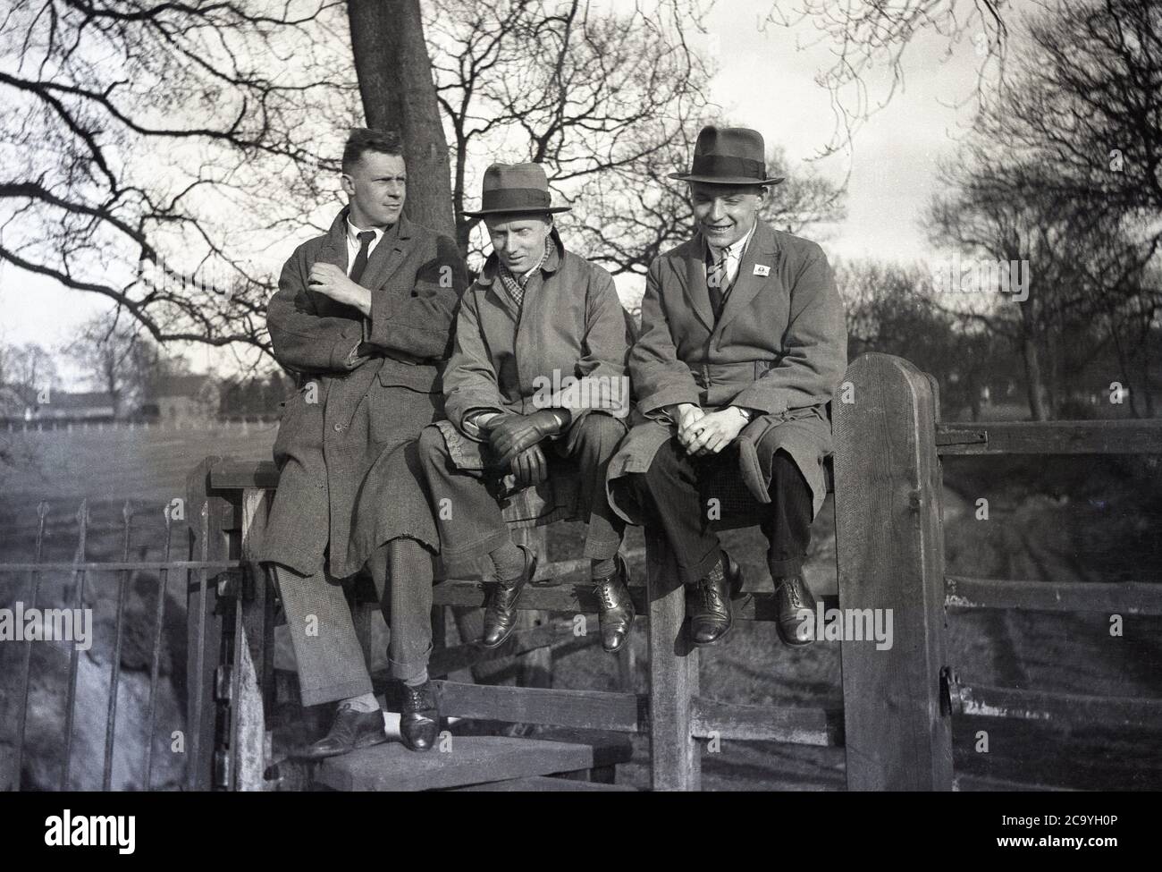 1940s/50s, historical, three smartly dressed, stylish men wearing overcoats and two with smart trilby hats, sitting together on a wooden fence by a park or possibly a school playing field, perhaps having been to a country horse racing meeting and thinking of what might have been if their bets had won, England, UK. A trilby is a narrow-brimmed hat, sometimes called a 'brown trilby' in Britain and frequently seen at horse race meetings. The name comes from the stage adaptation of George du Maurier's 1894 novel, Trilby. Stock Photo
