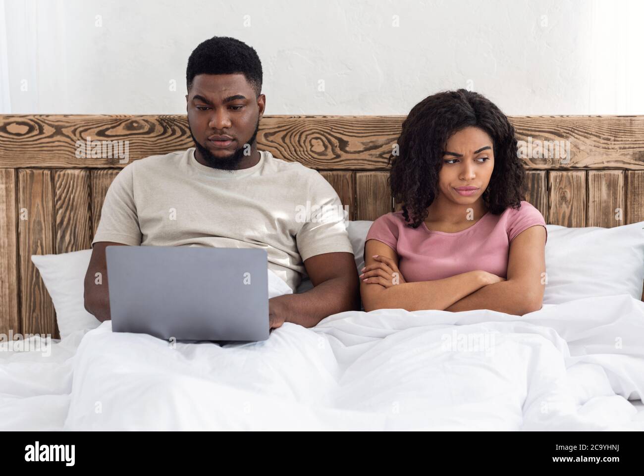 Addicted black man using laptop in bed, woman looking angry Stock Photo