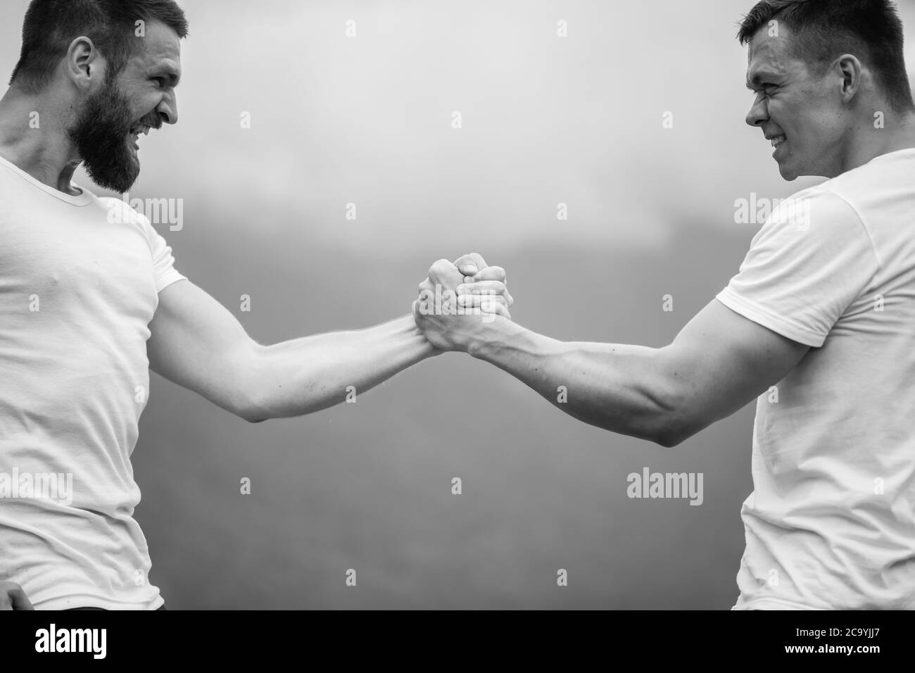 Strong male bodybuilders in white t-shirts greeting each other in wrestling manner outdoor over foggy mountain landscape. Rivalry, challenge, strength Stock Photo