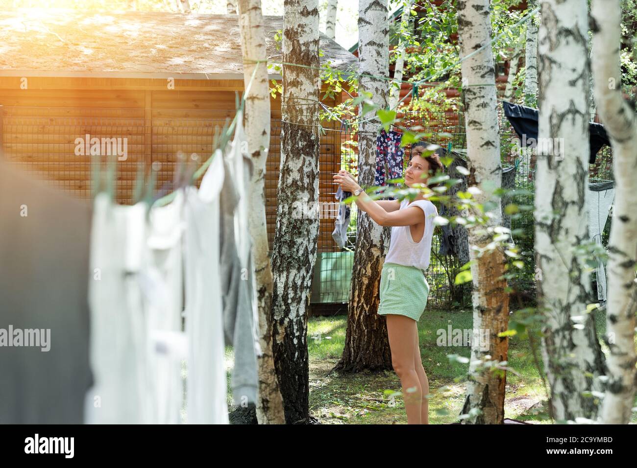 Candid real life portrait of young adult beautiful attractive caucasian woman hanging up fresh washed family clothes on birch tree clothesline with Stock Photo