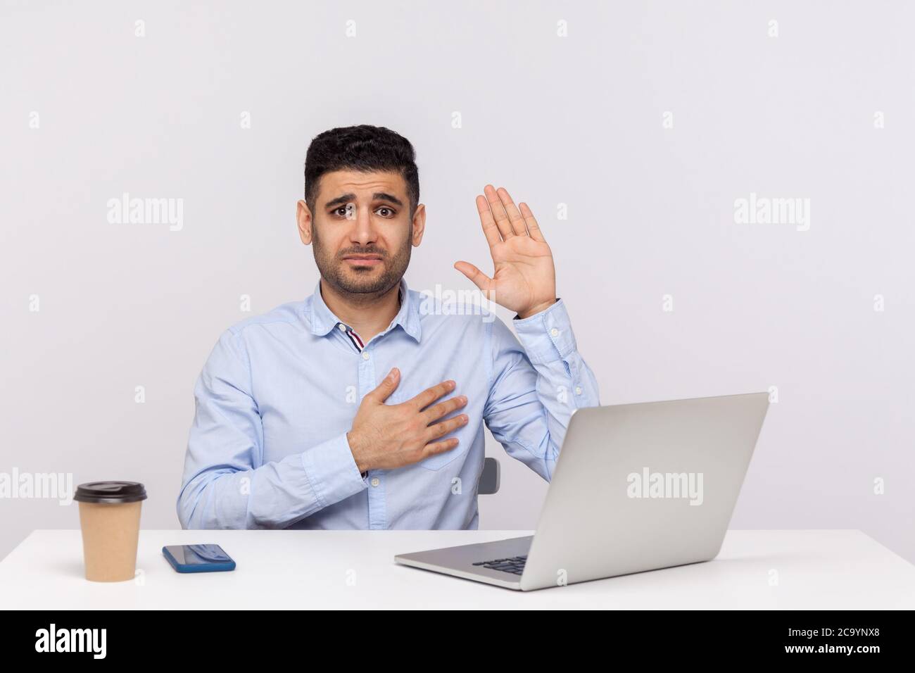 I swear! Honest businessman sitting office workplace with laptop on desk, raising hand, touching chest and making sincere promise, looking trustworthy Stock Photo