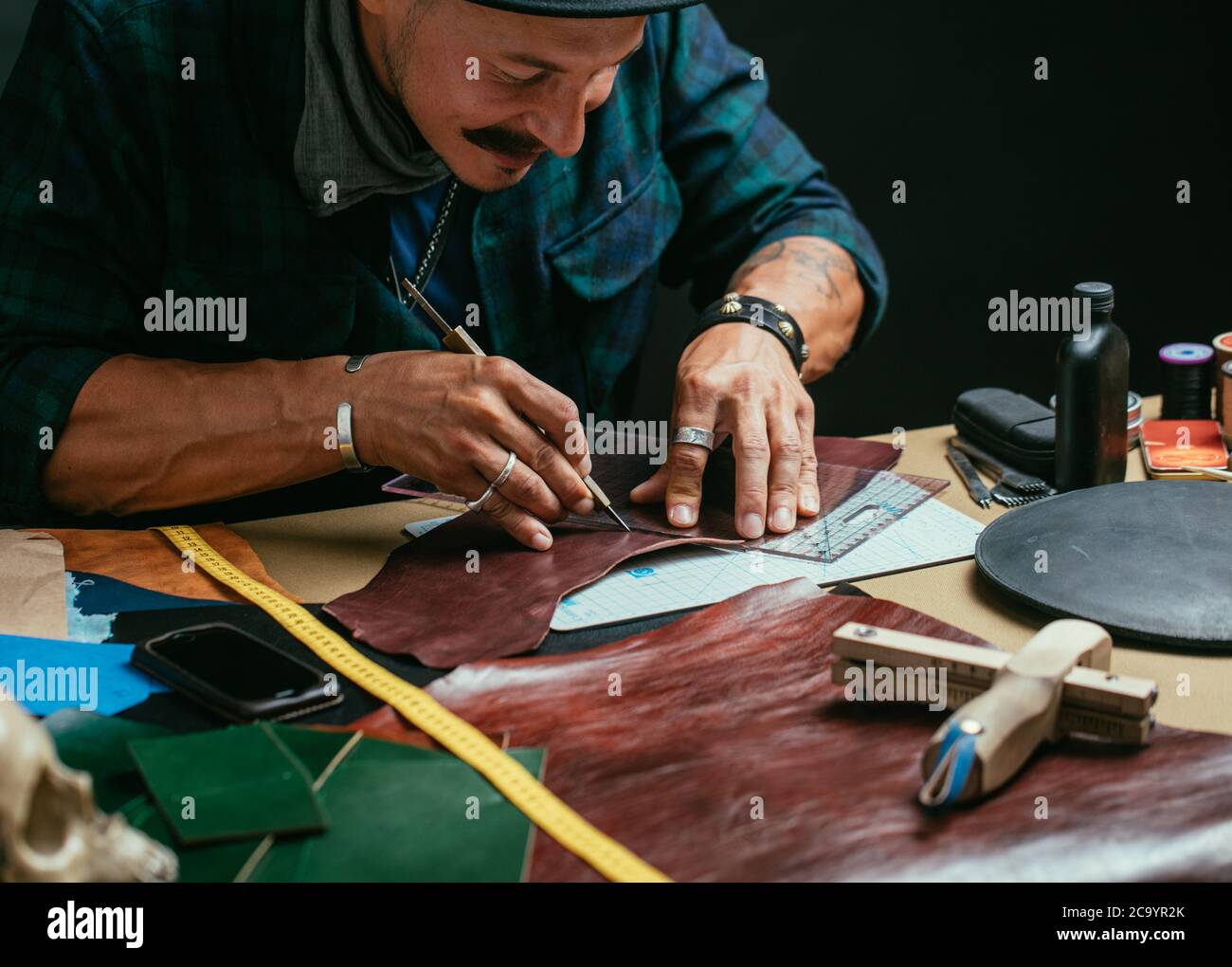 Mid aged caucasian stylish shoemaker wearing black hat measuring leather at footwear workshop against black wall. Small Business and craftspersons Con Stock Photo