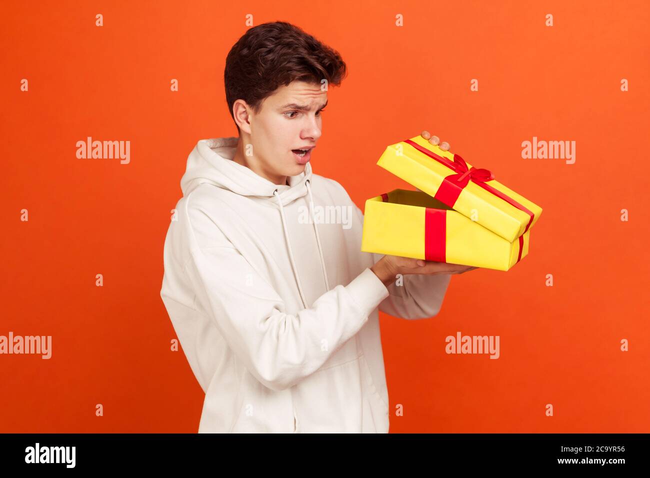 Shocked young man in casual white sweatshirt with hood glancing inside present box, disappointed witt gift, unjustified expectations. Indoor studio sh Stock Photo