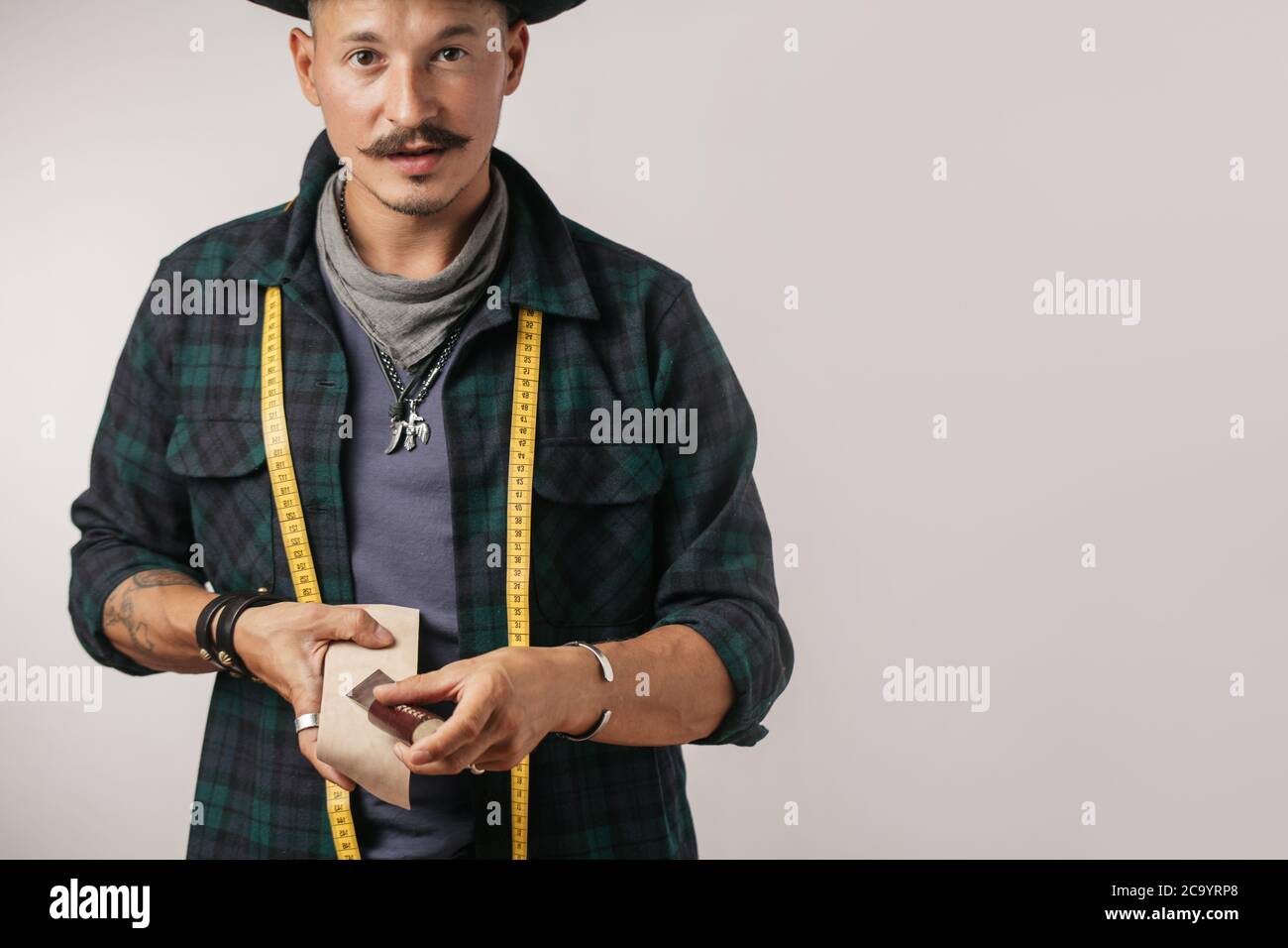 Studio portrait of smiling caucasian mid-aged cobbler wearing trendy hat with measuring tape slapping sole on man s leather boot. Stock Photo