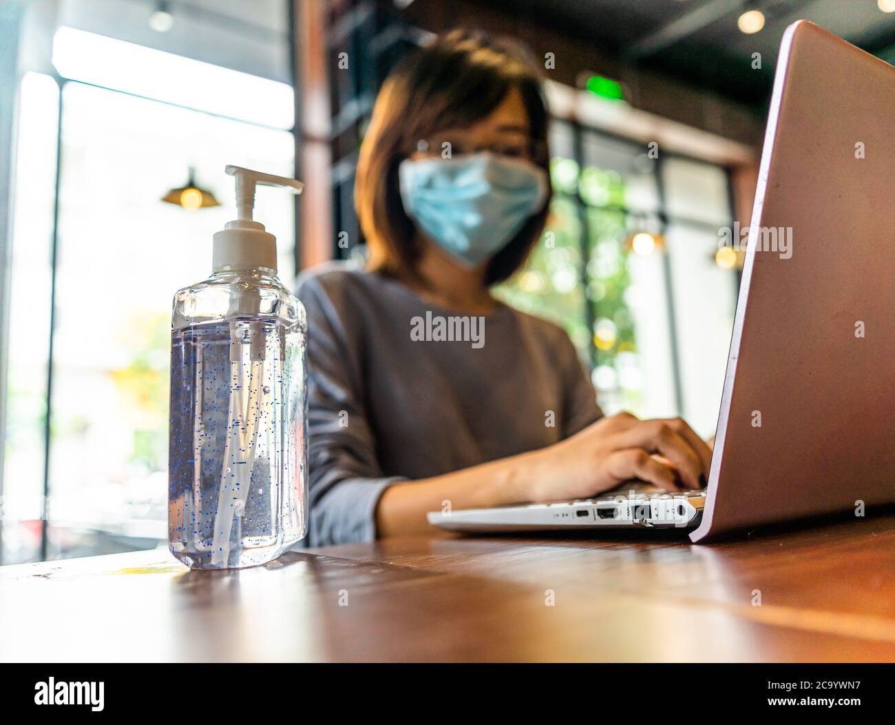Young asian woman using laptop computer working at home with alcohol gel in quarantine for coronavirus wearing protective mask with social distancing Stock Photo