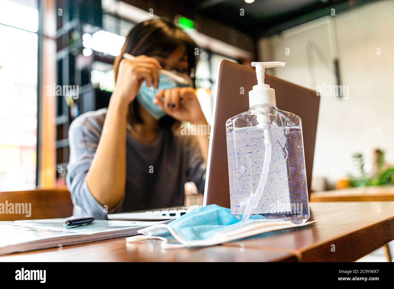 Young asian woman using laptop computer working at home with alcohol gel in quarantine for coronavirus wearing protective mask with social distancing Stock Photo