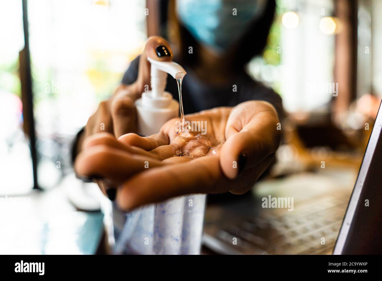 Young woman washing hands with alcohol gel in quarantine for coronavirus wearing protective mask with social distancing and using laptop computer work Stock Photo