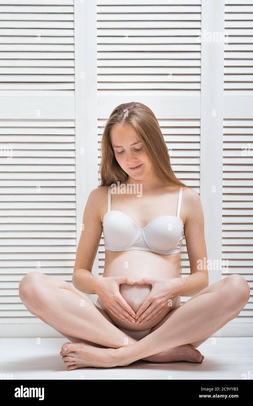 Young beautiful pregnant woman sitting in pose of Lotus and put hands in heart shape on stomach. Vertical frame. Stock Photo