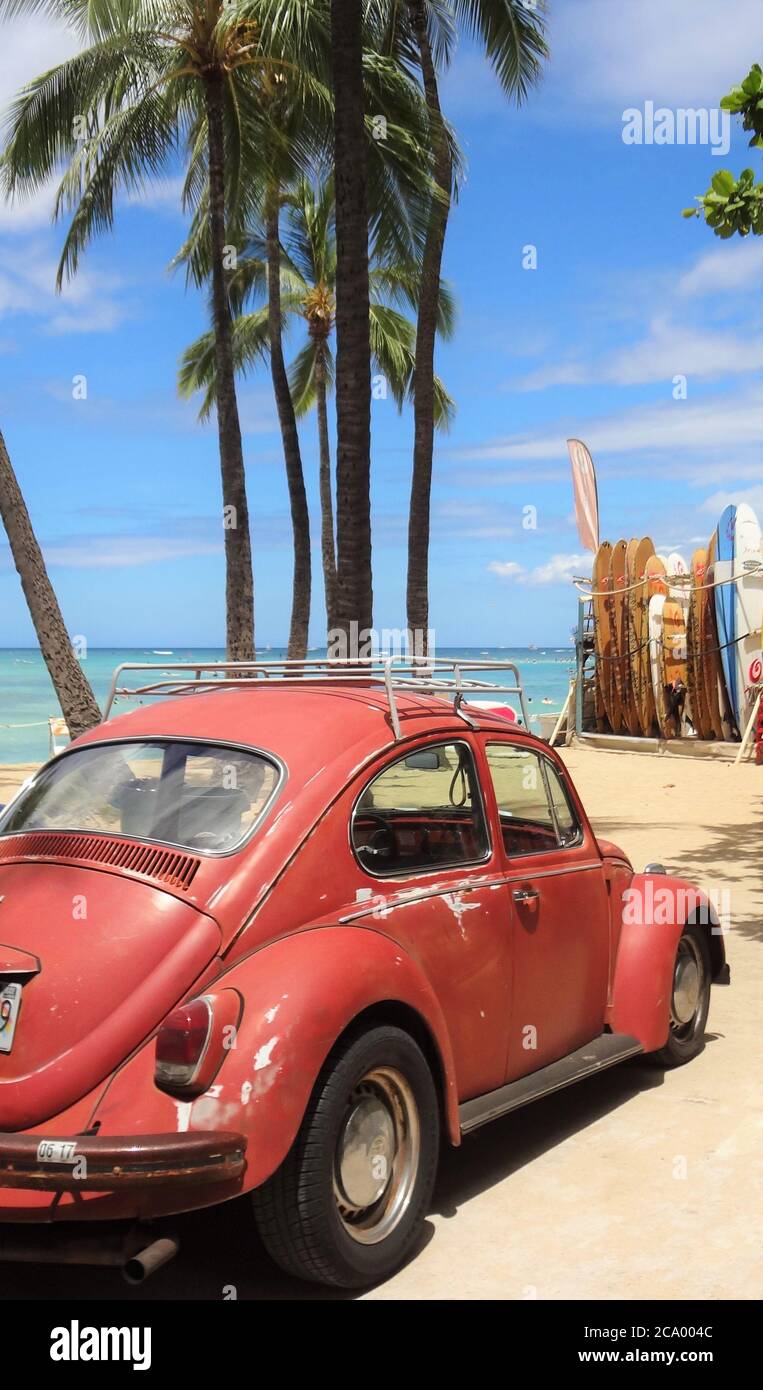 A red VW Beetle on Waikiki beach with palm trees and surfboards against a blue sky Stock Photo