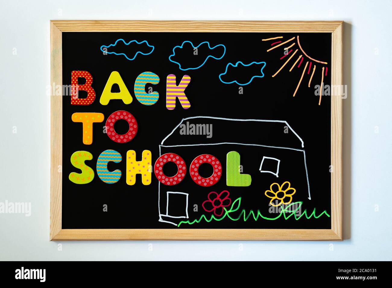 Blackboard with text Back to school of colorful wooden letters and chalk drawing of schoolhouse, grass, flowers, sun, and clouds. Words Back to school Stock Photo