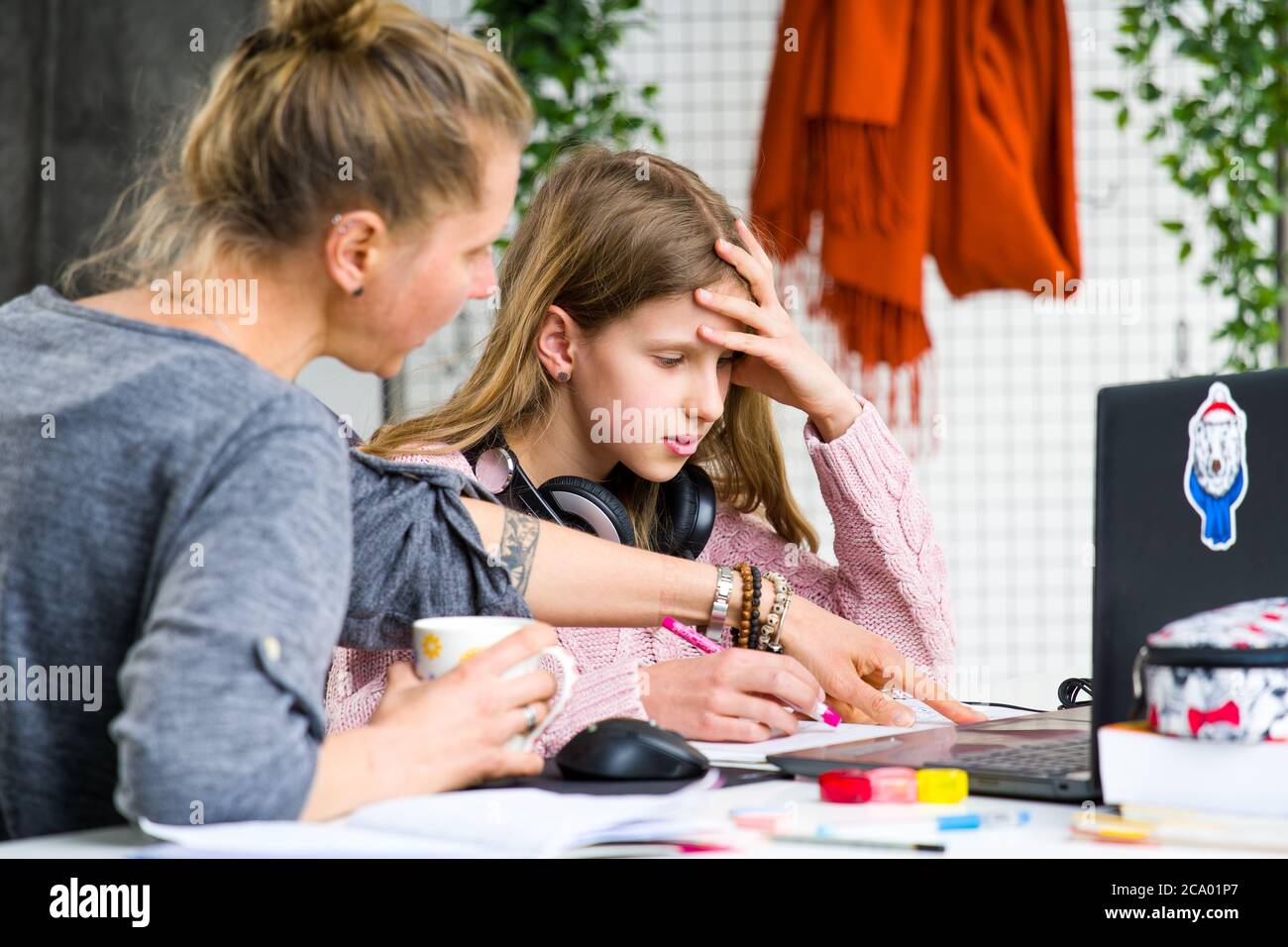 Teenage, blond girl in pink sweater is confused and asking her mother for help while doing her lessons. Mother is drinking a tea and trying to help. Stock Photo