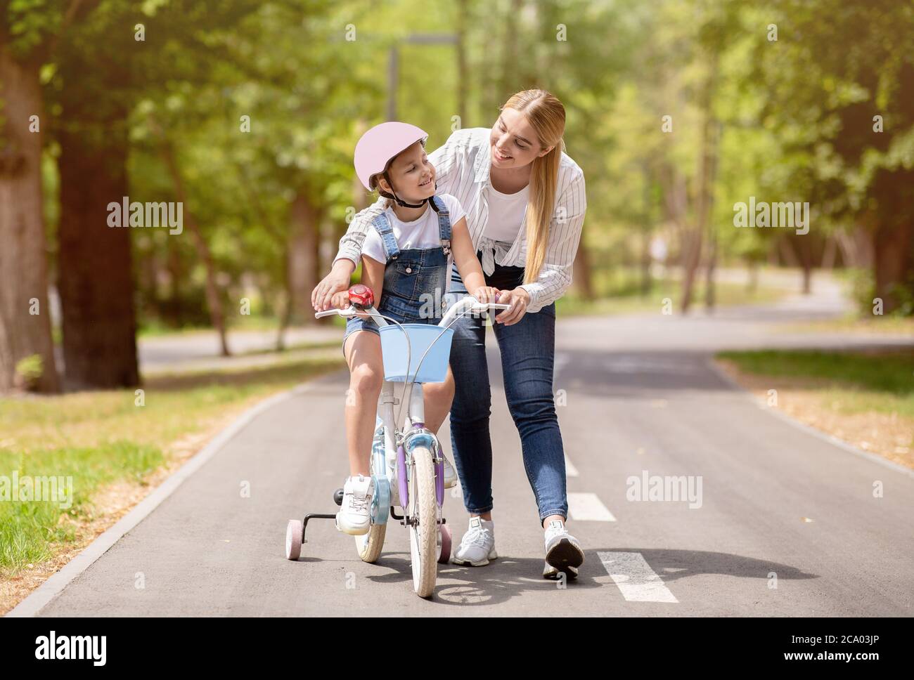 Mom Teaching Little Daughter To Ride Bicycle With Stabilisers Outdoors