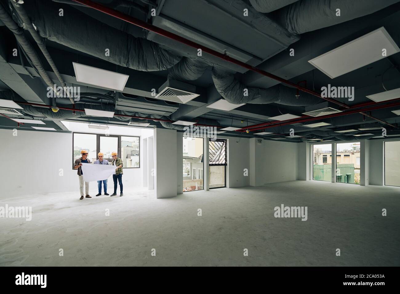Group of young construction engineers in hardhats checking blueprint of building they are working on Stock Photo