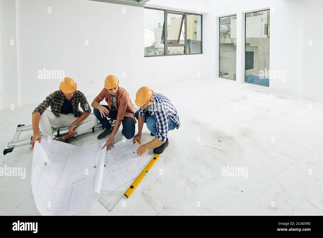 Team of young builders in hard hats checking blueprints before starting renovation process in apartment of client Stock Photo