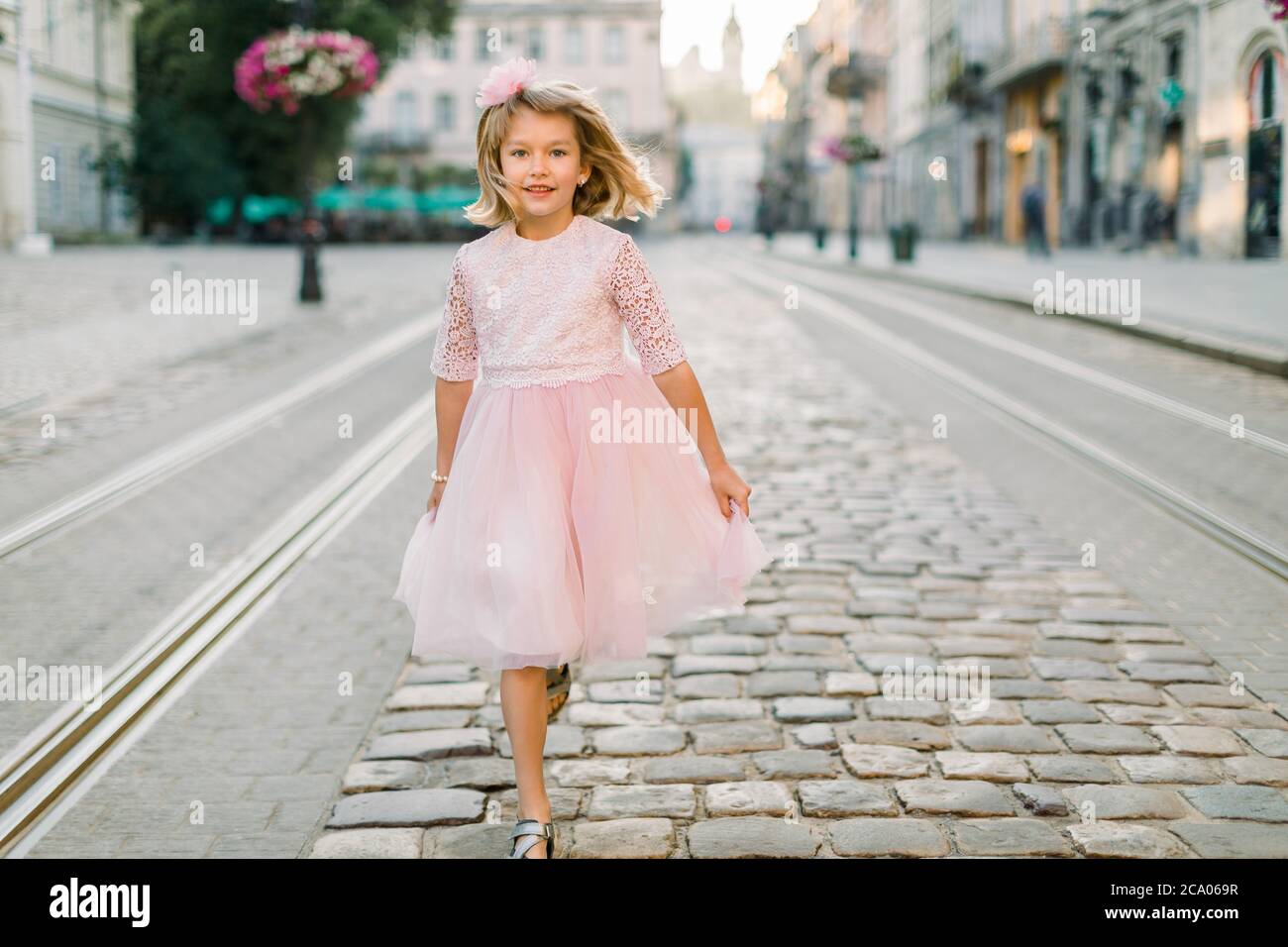 Cute blond little girl walks along the street of the old European city in her beautiful pink dress. Front view. Copy space Stock Photo