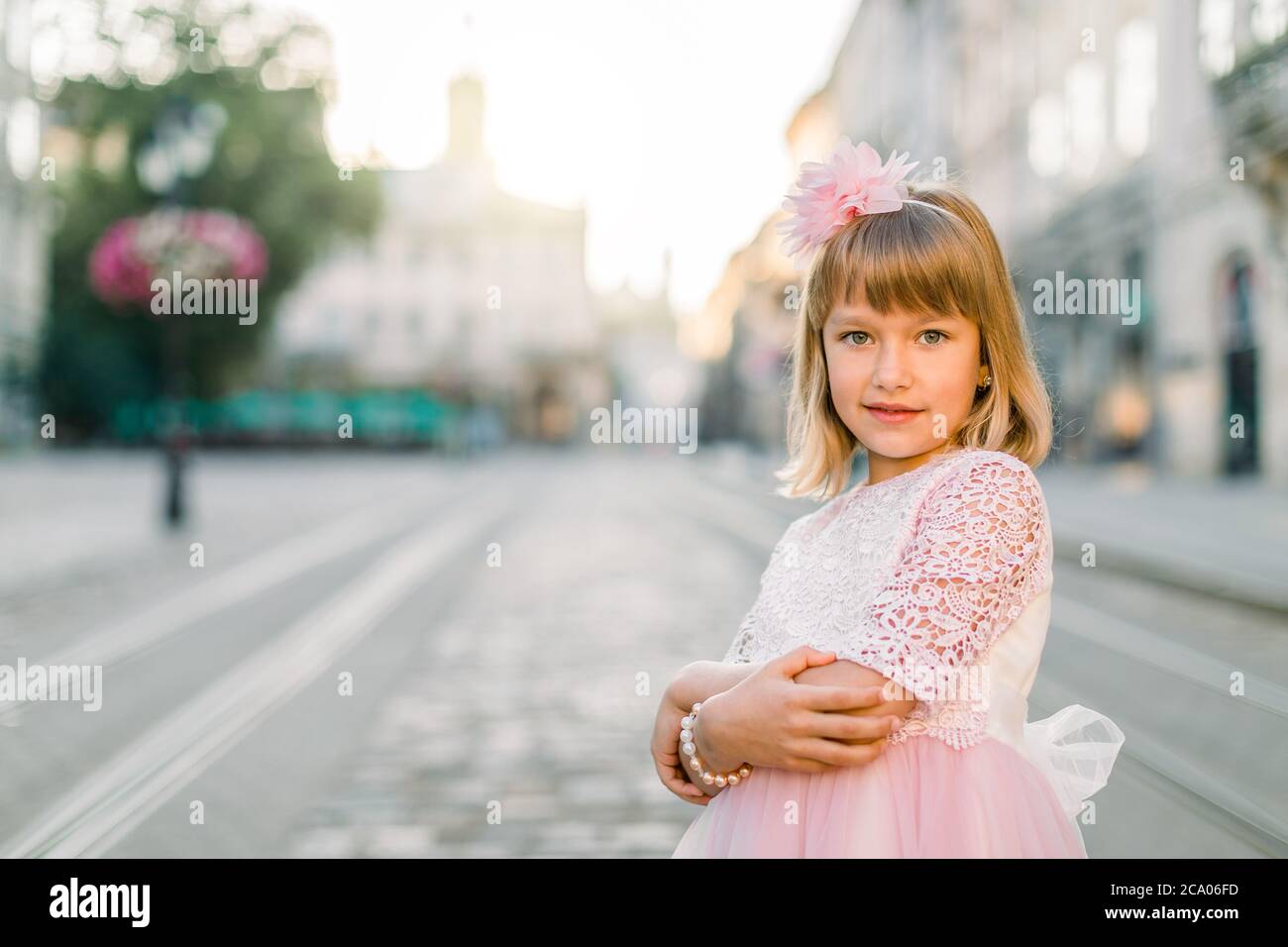 Outdoor close up shot of little girl in a pink dress with a pink flower in her hair, posing to camera in front of old city street. Portrait of a girl Stock Photo