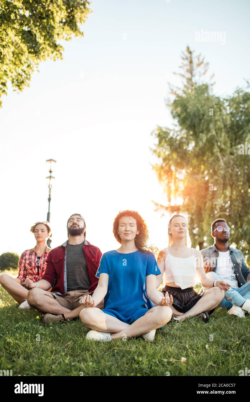Time for yoga, Attractive young multicultural friends meditating in yoga lotus pose while resting after a long trip in Botanical garden. Multiethnic p Stock Photo