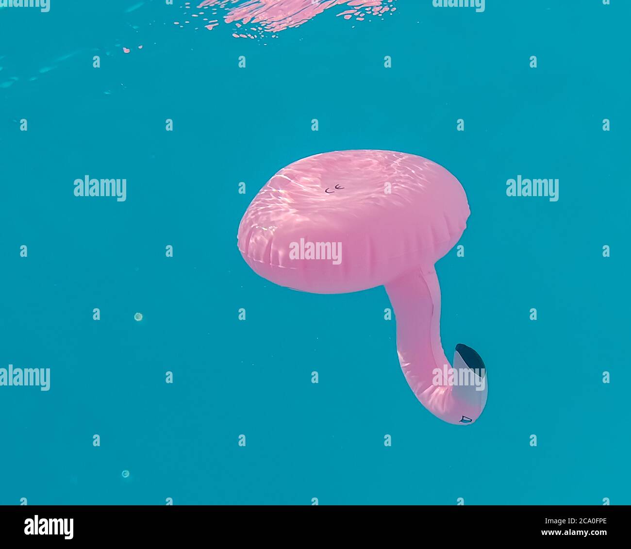 Inflatable pink flamingo can holder with a CE-mark is floating upside down in turquoise water, with reflections visible on the surface. Stock Photo