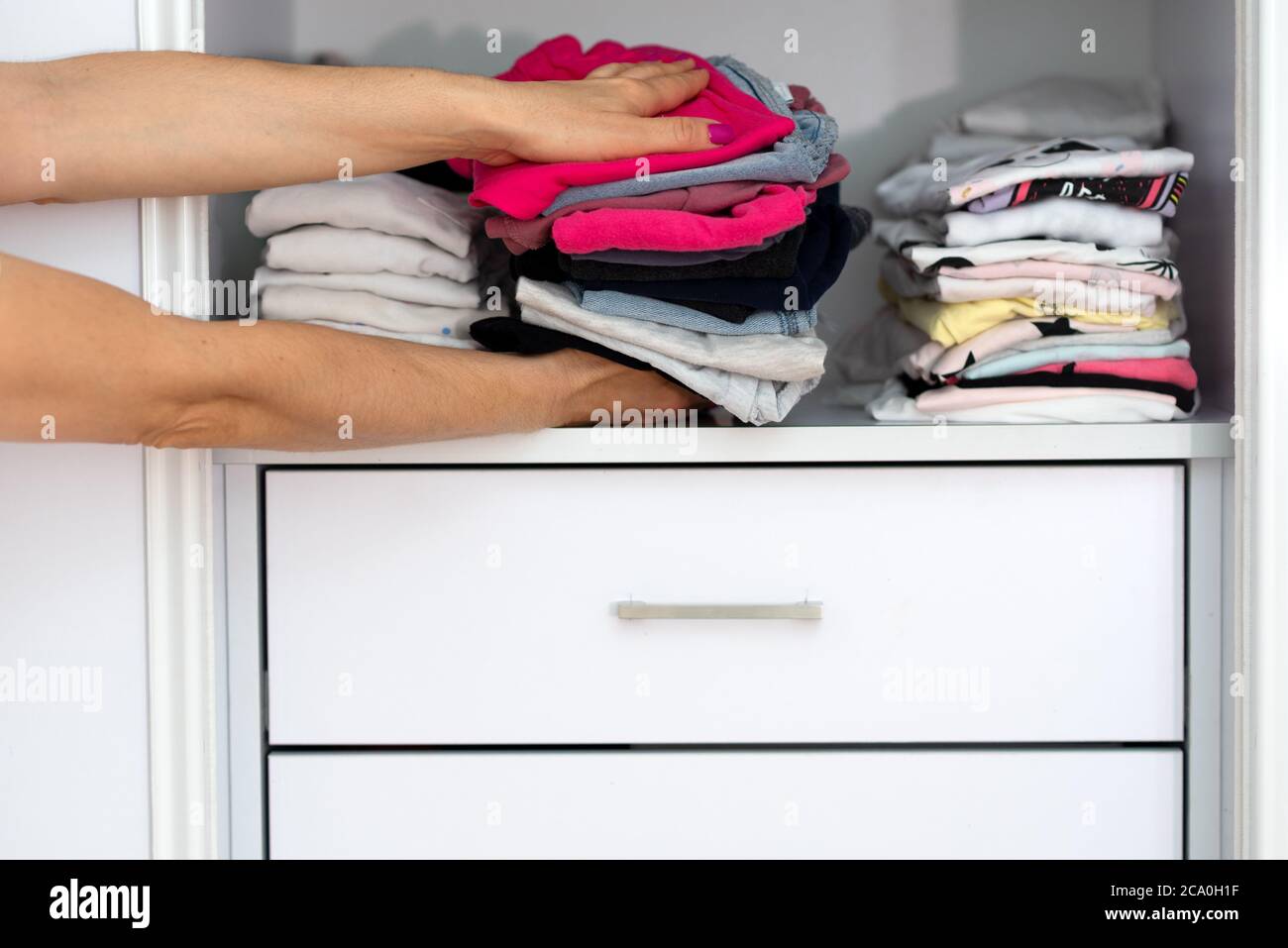 Mom picking out kid outfit at home to send her children back to school. New colored clothing stacked in a pile close-up in white wardrobe. Woman folding clothes on a shelf in a closet. Stock Photo