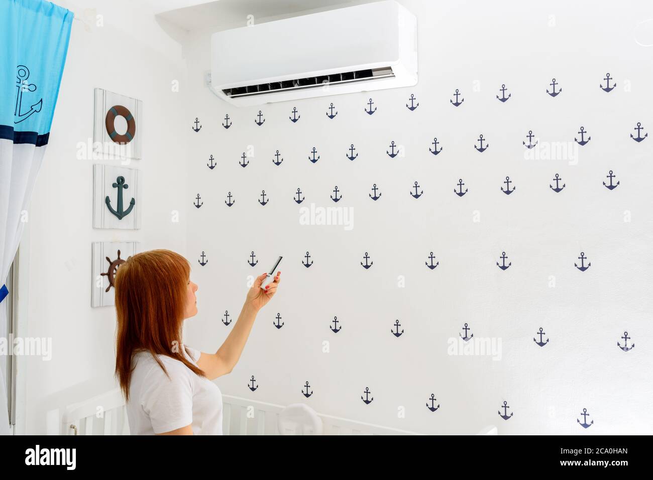 Portrait Of A Happy Woman Holding Remote Control In Front Of Air Conditioner At Home in childrens room.Air conditioner inside the cute baby boys room.With hot weather,is a good solution for family. Stock Photo