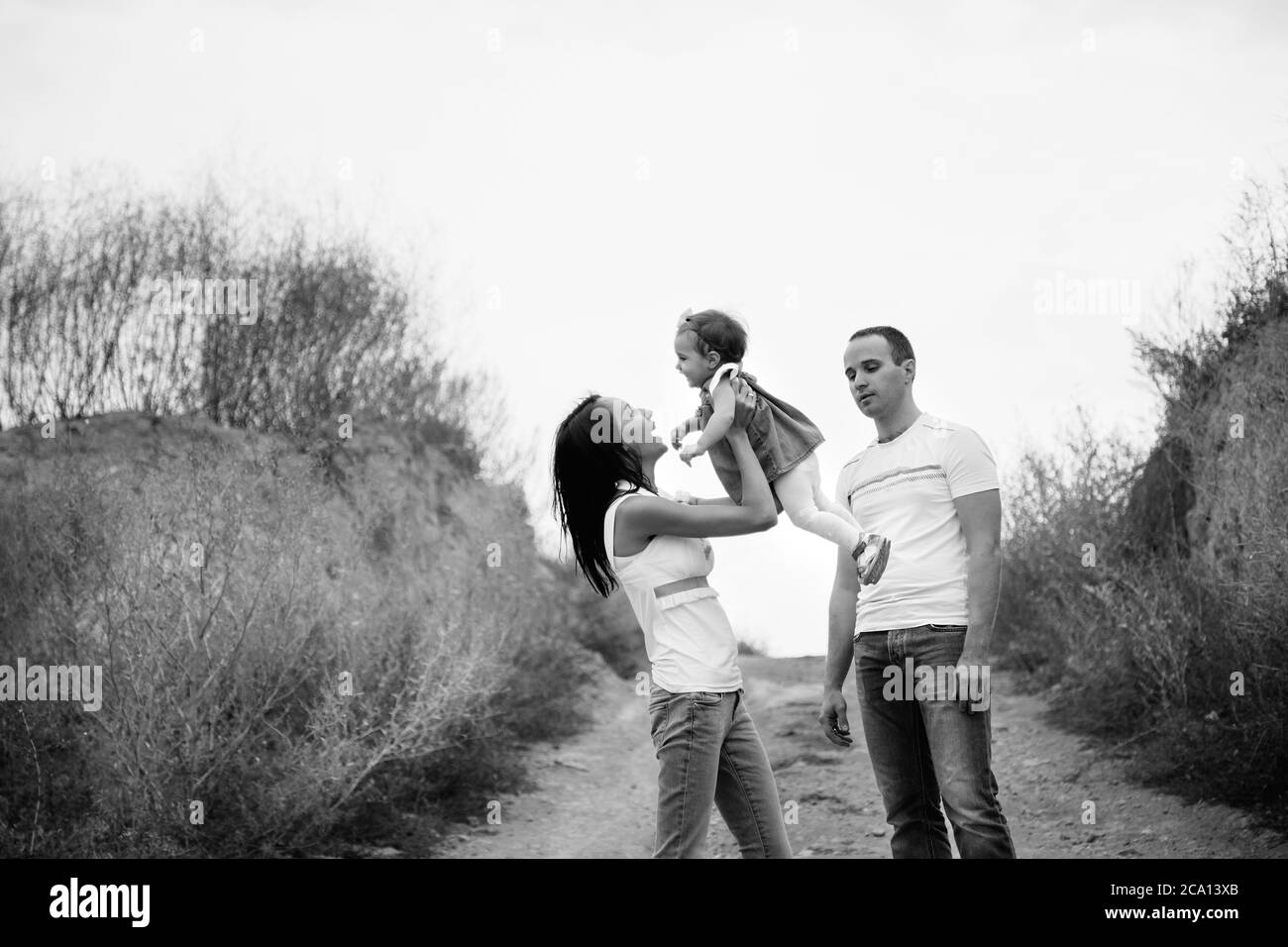 Happy young family in white t-shirts and blue jeans with a small daughter in dress hugging, outdoors background Stock Photo