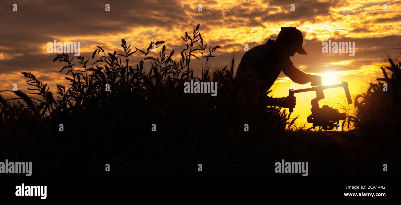 Video Operator with Modern Camera Installed on a Gimbal Walking Along Countryside During Scenic Sunset. Panoramic Silhouette Concept. Stock Photo