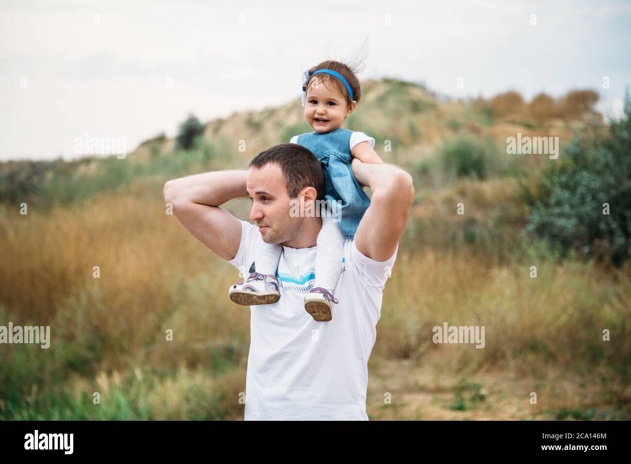 Close up of small baby girl in blue jeans dress sitting on father's shoulders Stock Photo
