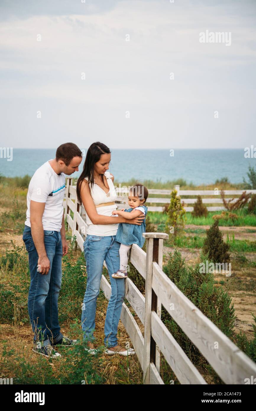 Close up of small baby girl in blue jeans dress sitting on a fence with mother and father near Stock Photo