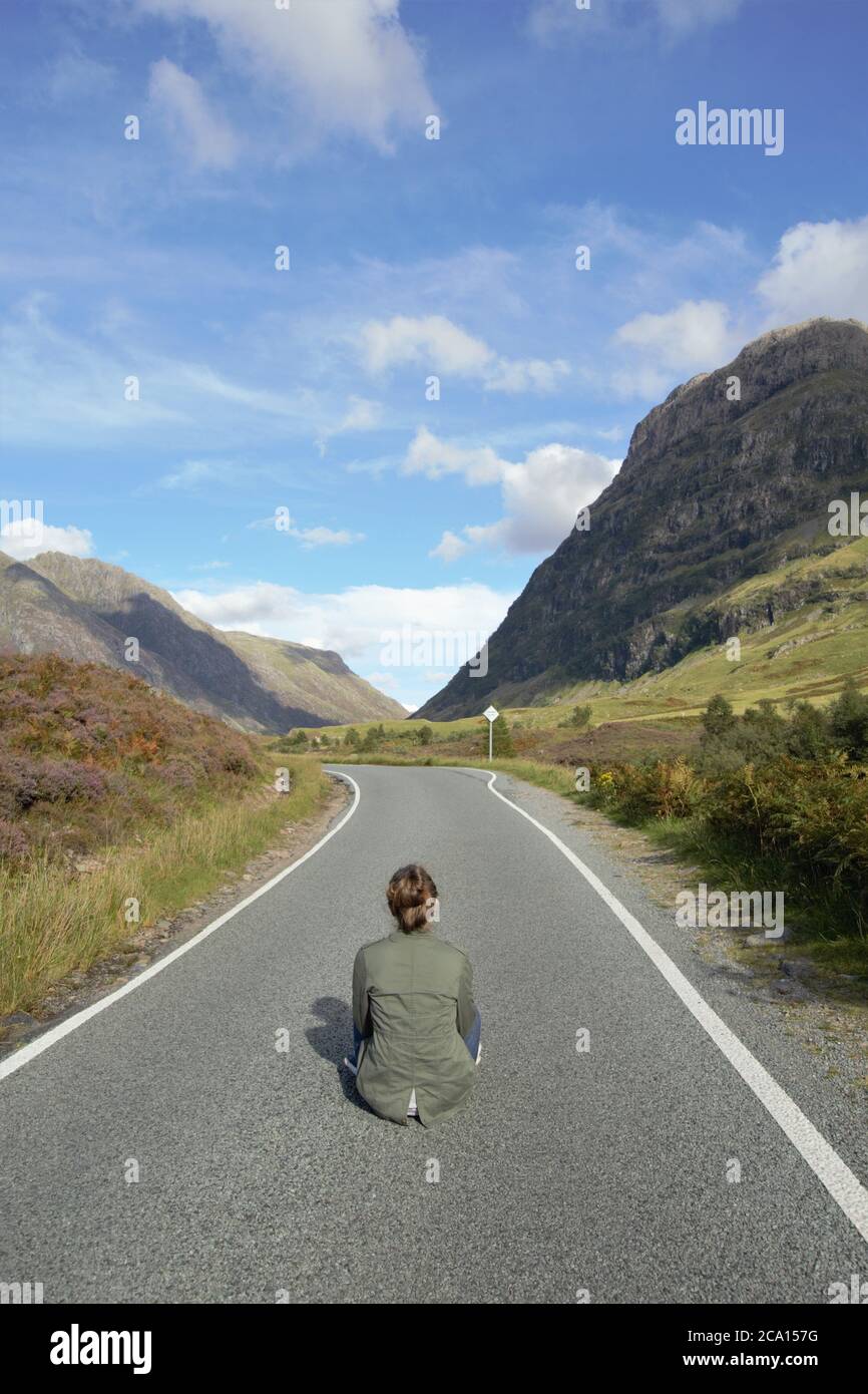 A young girl sitting on the asphalt road enjoying the magnificent view and freedom in Glencoe in Scottish Highlands in Summer, Scotland, Great Britain Stock Photo
