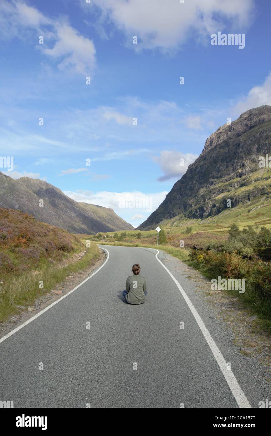 A young girl sitting on the asphalt road enjoying the magnificent view and freedom in Glencoe in Scottish Highlands in Summer, Scotland, Great Britain Stock Photo