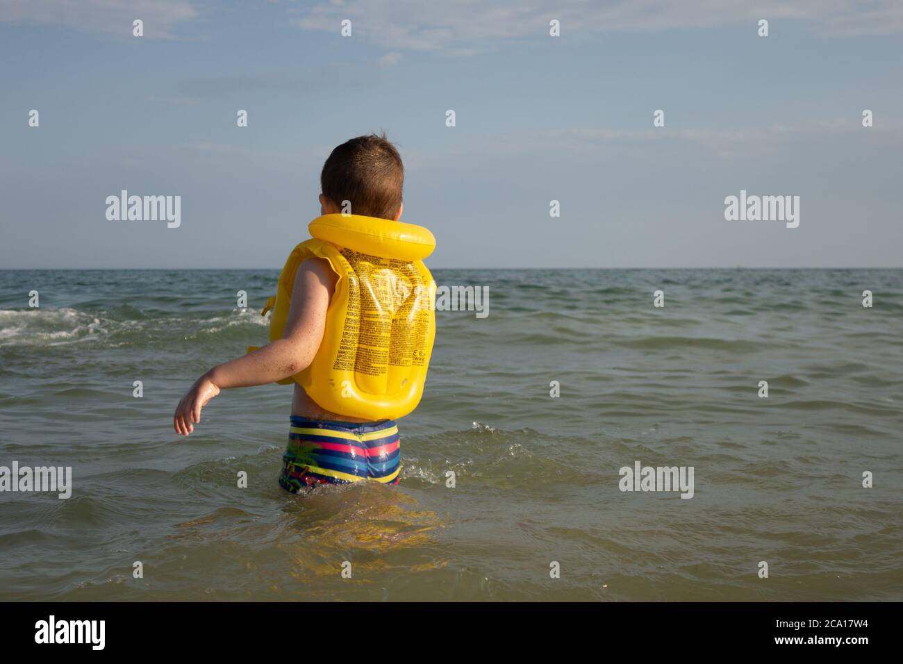 A 5-year-old boy in a yellow life jacket and rainbow-colored shorts enters the sea. Stock Photo