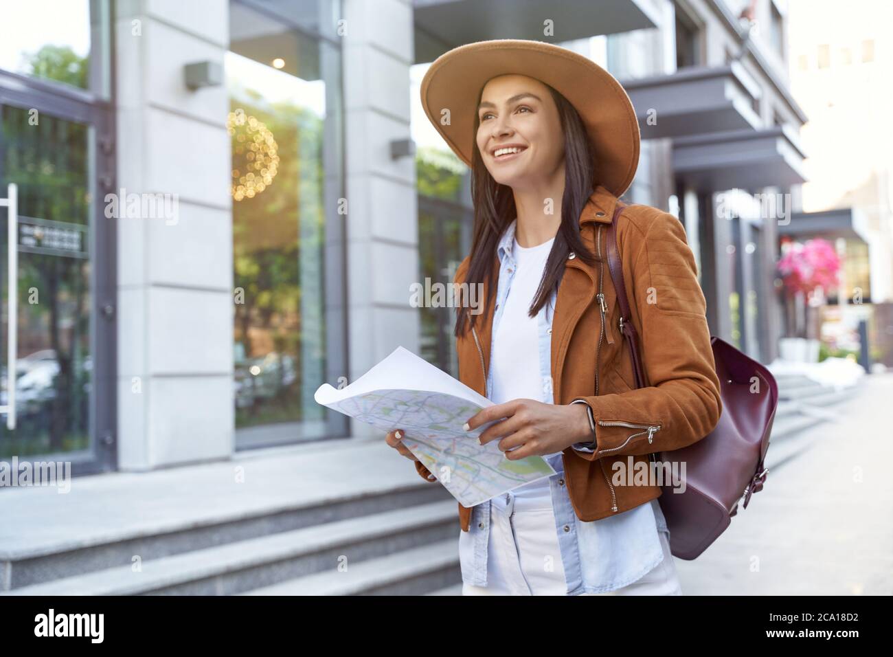 Traveling abroad. Young happy female traveler or tourist wearing hat and backpack holding map and smiling while walking on city street. Travel, tourism, vacation Stock Photo