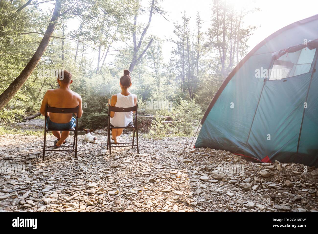 Relaxed couple sitting outside tent while camping nearby mountain river on a sunny day. Woman holding coffee talking with man. Two young caucasian people enjoying camping together. Stock Photo