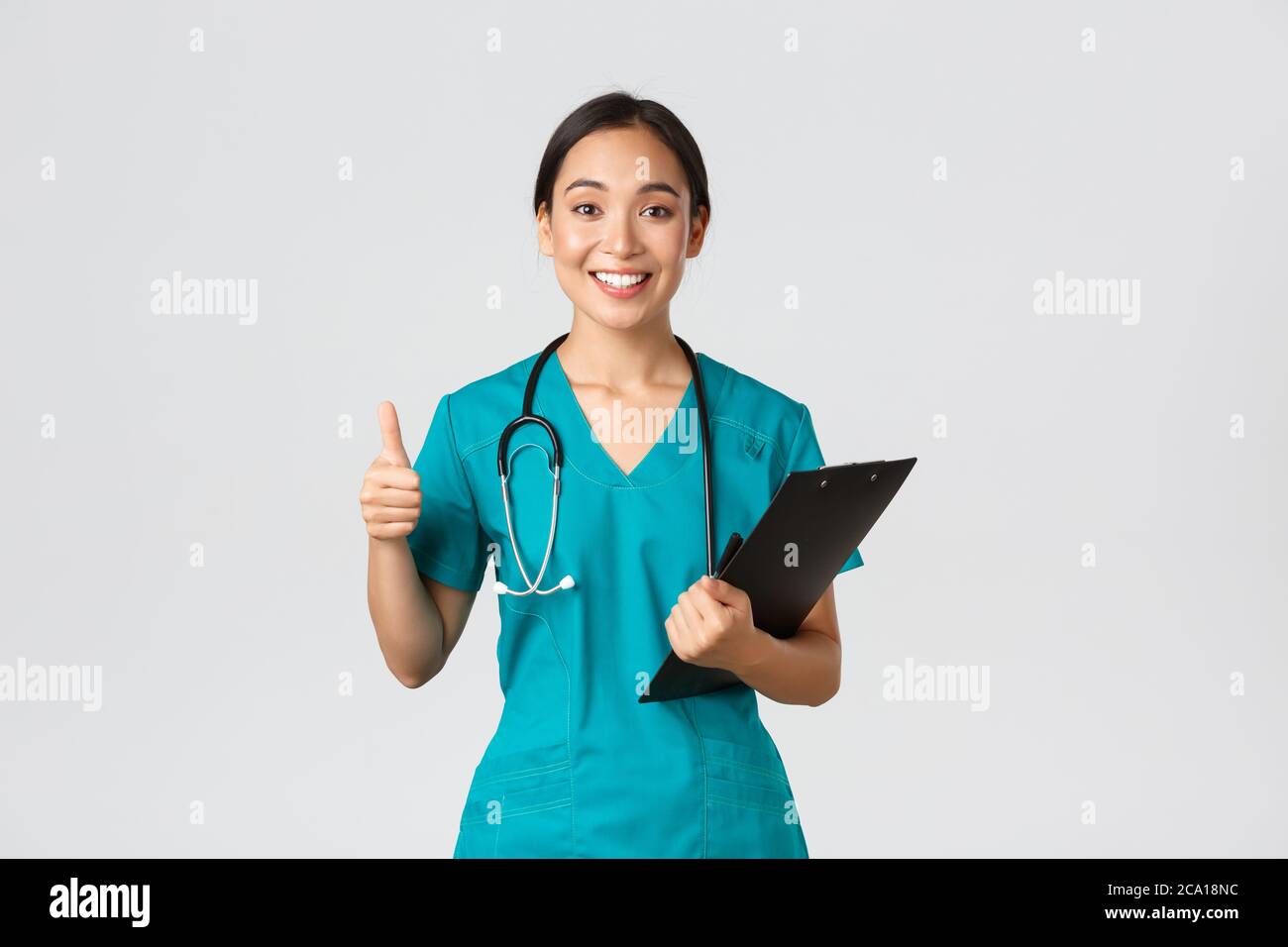 Healthcare workers, preventing virus, quarantine campaign concept. Smiling pretty asian doctor, female intern in scrubs show thumb-up, holding Stock Photo