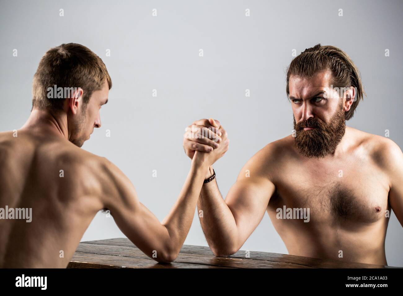 Arm wrestling. Heavily muscled bearded man arm wrestling a puny weak man. Arms wrestling thin hand, big strong arm in studio. Two man's hands clasped Stock Photo