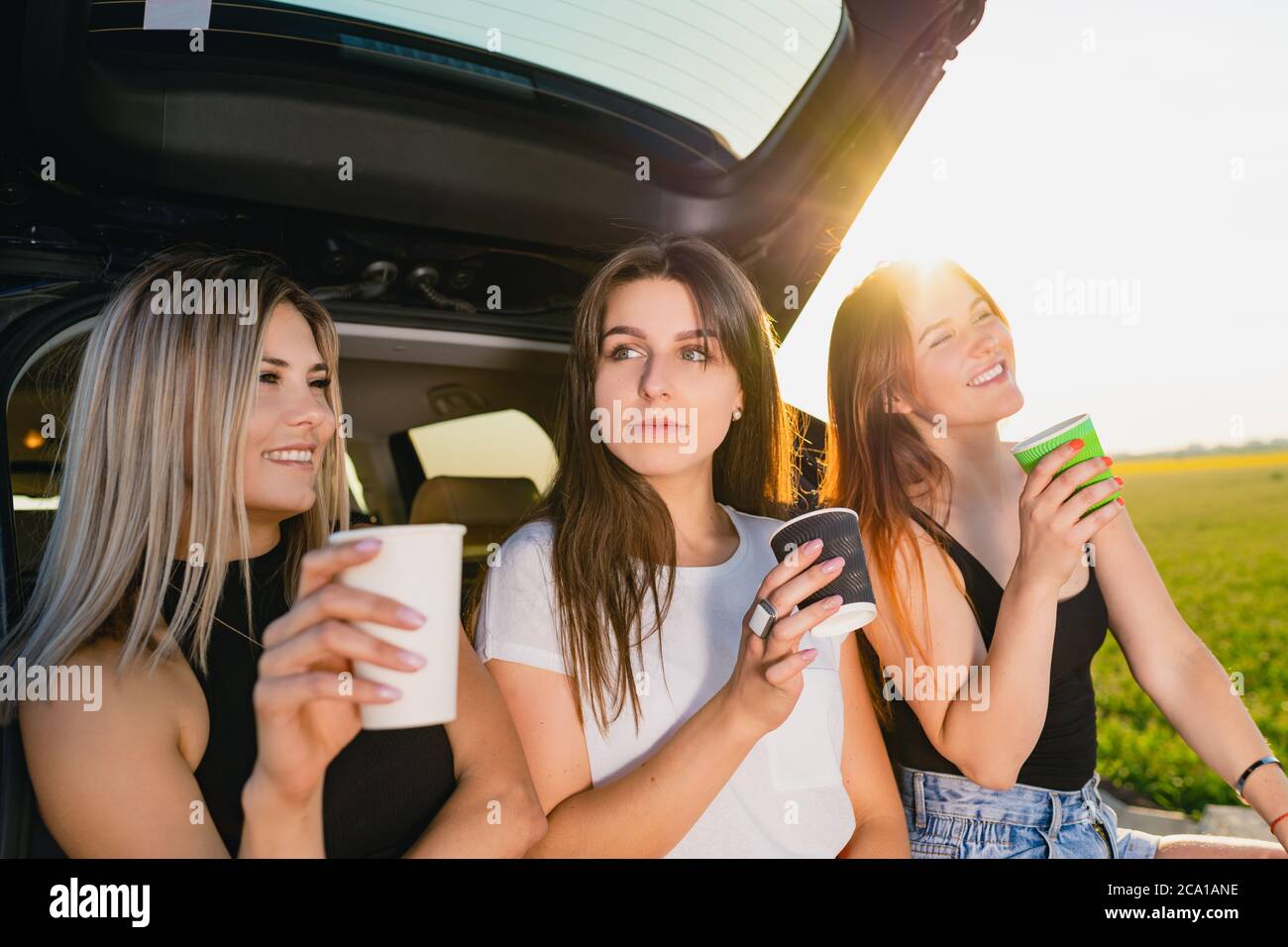 Three girls have coffee break at car trunk. Happy road trip smiling female travelers, relax at vehicle back with coffee cups take away at sunlight. Stock Photo