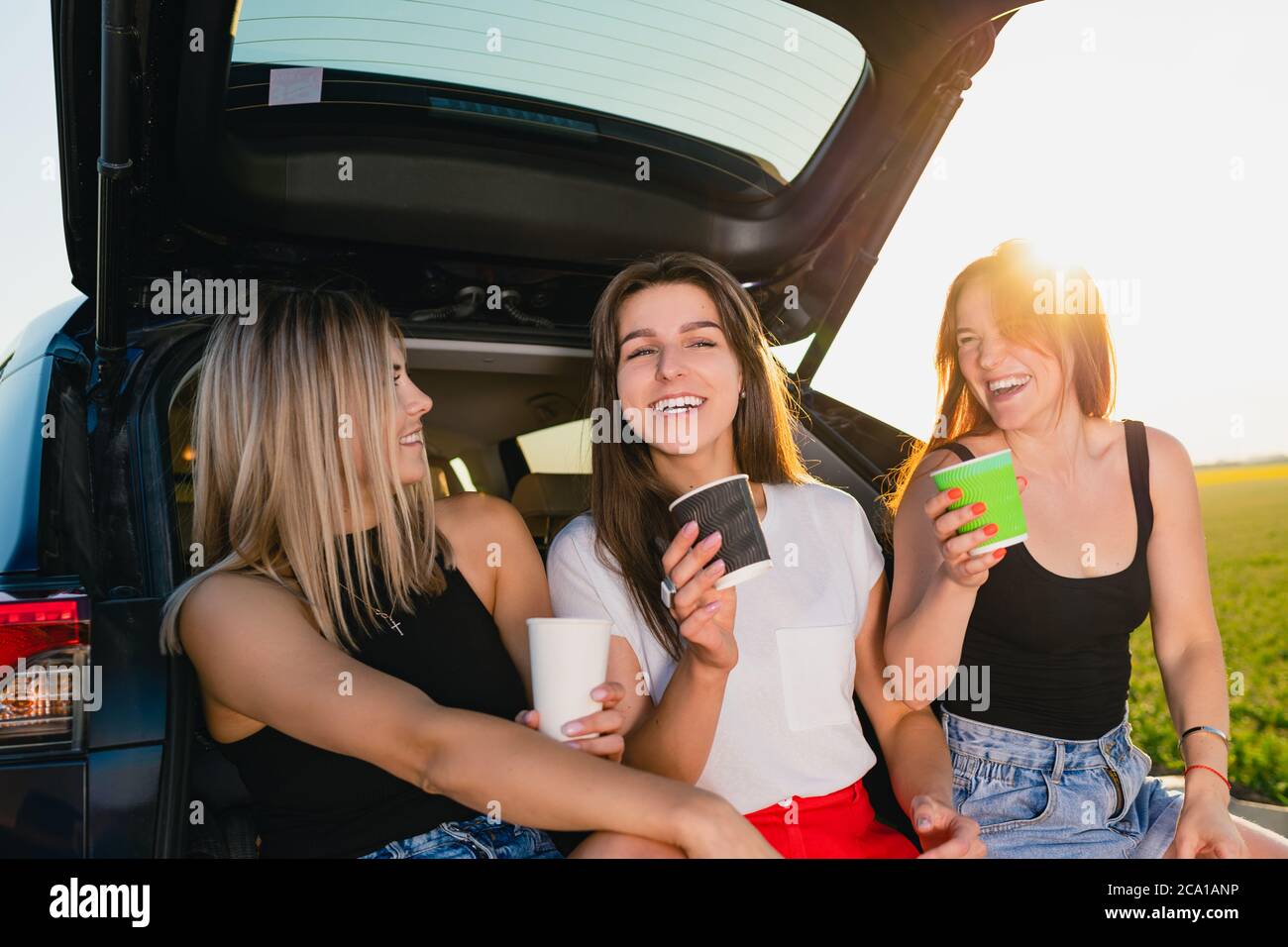 Happy road trip of three smiling women, relax at vehicle back with coffee cups take away at sunlight. Stock Photo