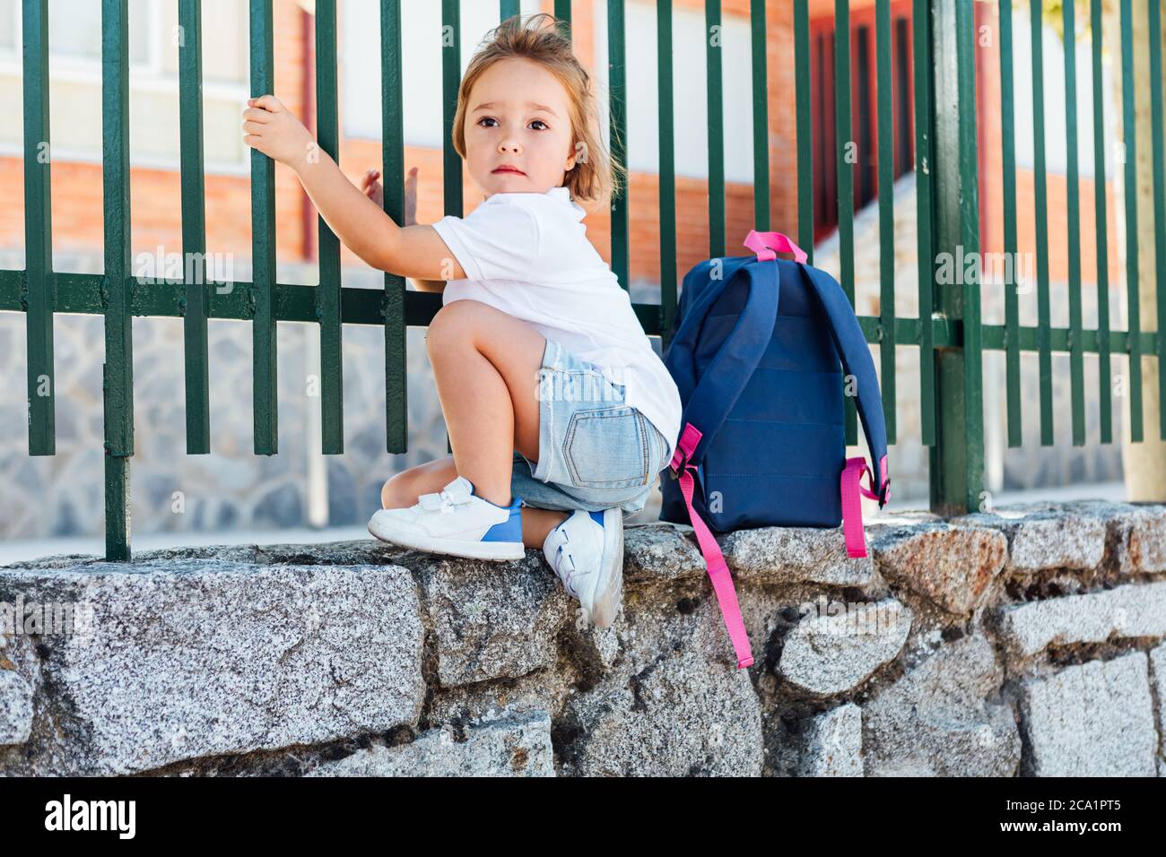 A blond-haired girl carrying a blue backpack on the school fence. School concept Stock Photo