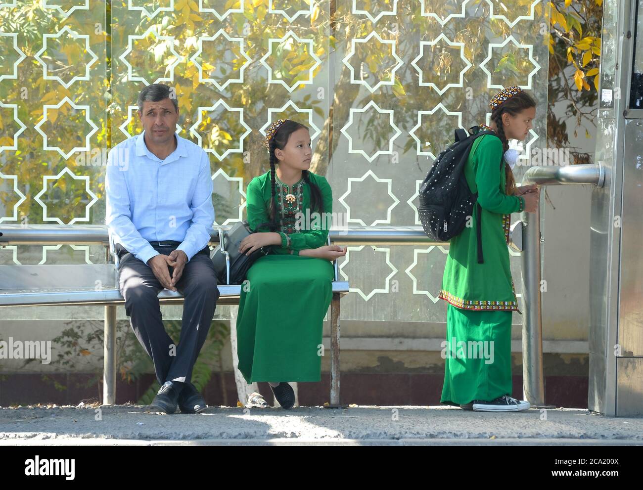 Young turkmen girls wearing the Turkmenistan national green dress used as school uniform accompanied by thoughtful man in a bus stop in Ashgabat. Stock Photo