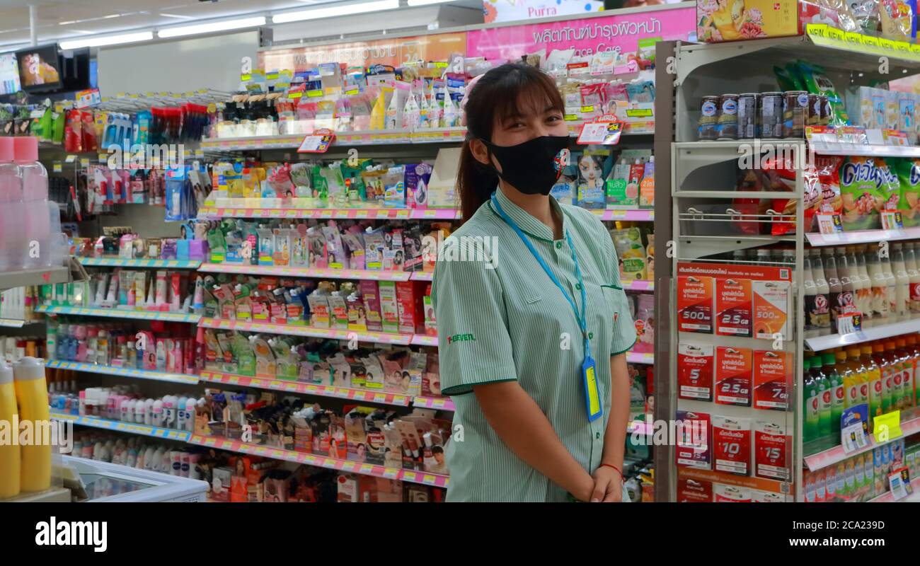 Bangkok, Thailand- August 3, 2020: Smiling saleswoman wearing cloth face mask standing in front of shelves near entrance of convenience store Stock Photo