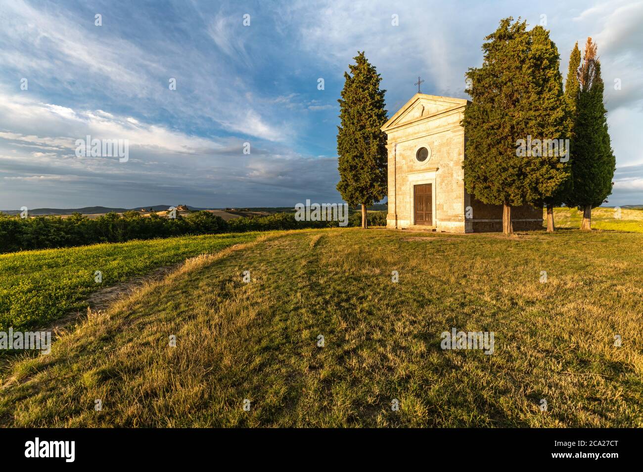 Wide angle view of the iconic Tuscanian hilltop chapel of Vitaleta, surrounded by cypresses, under a summer blue sky with streaked clouds Stock Photo
