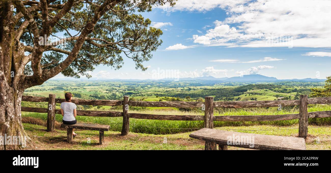 Lush farmland panoramic scene with a female seated on a stone bench in front of rustic hand-hewn wooden fence at Millaa Millaa Lookout in Queensland. Stock Photo