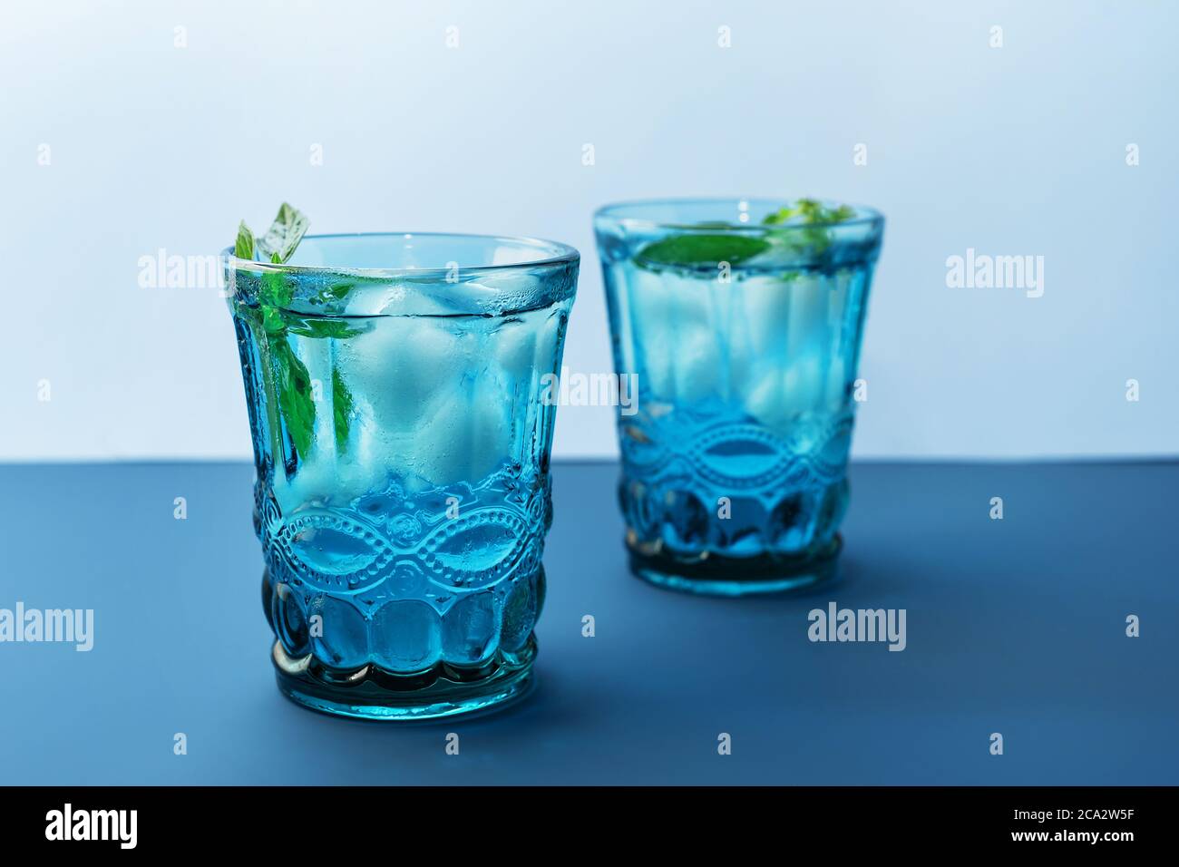 Two beautiful vintage blue glasses with cold drink and ice cubes, with fresh green mint leaves on deep blue and light blue background. Backdrop for yo Stock Photo