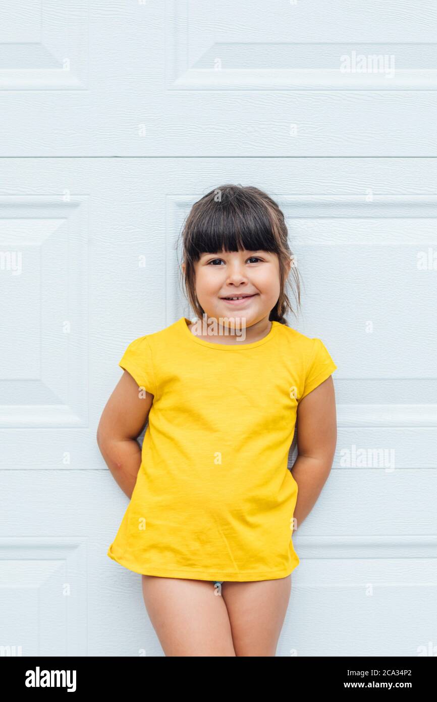 Adorable little girl with black hair wearing a yellow shirt leaning against white background. Stock Photo