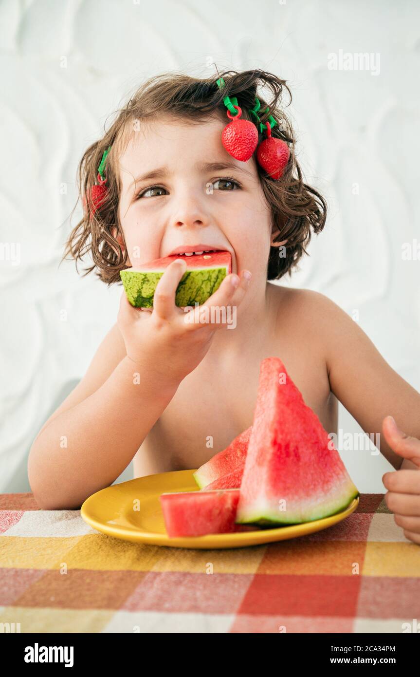 Hungry cute girl devours watermelon slices, refreshing summer dessert. Has strawberry hair clips acting being goofy funny. Stock Photo