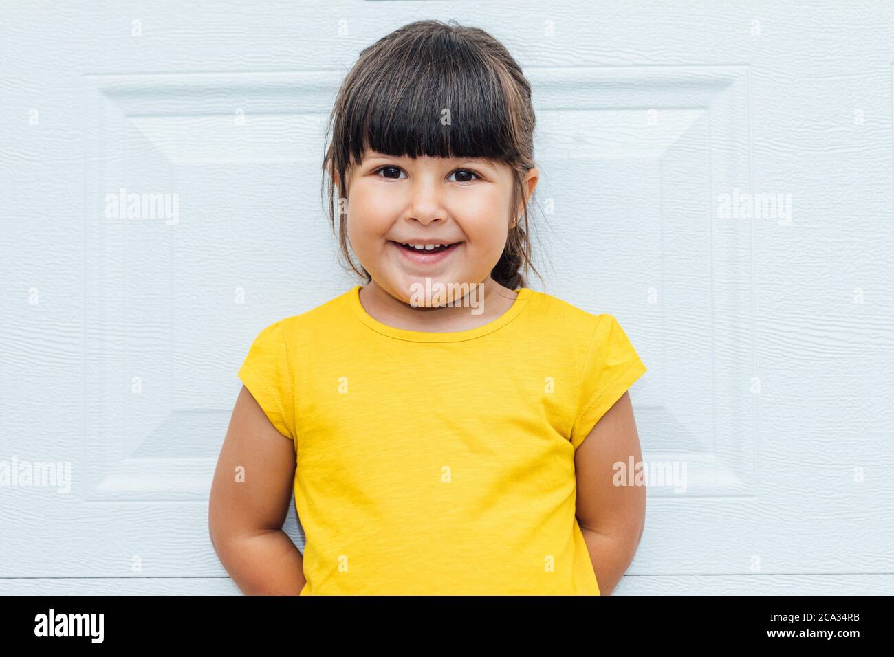 Adorable little girl with black hair wearing a yellow shirt leaning against white background. Stock Photo