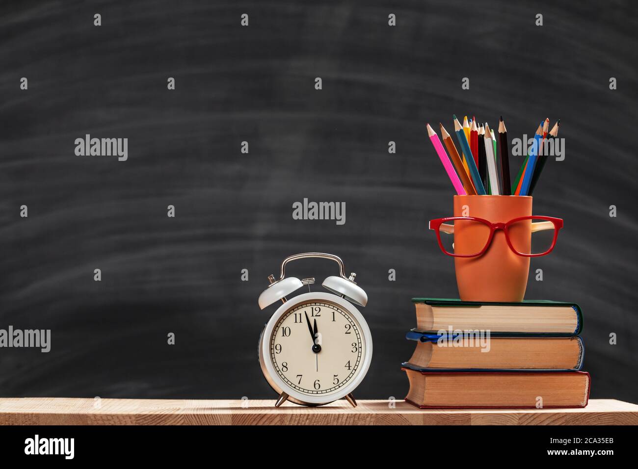 Student set. A stack of books, pencils, an alarm clock, glasses on the background of a school black board. Education concept. Copy space. Stock Photo