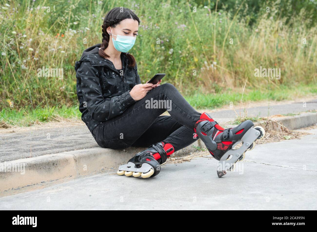 Woman in protective face mask, on roller skating pause, sitting on the street and using mobile phone during coronavirus pandemic outbreak. Urban Girl Stock Photo
