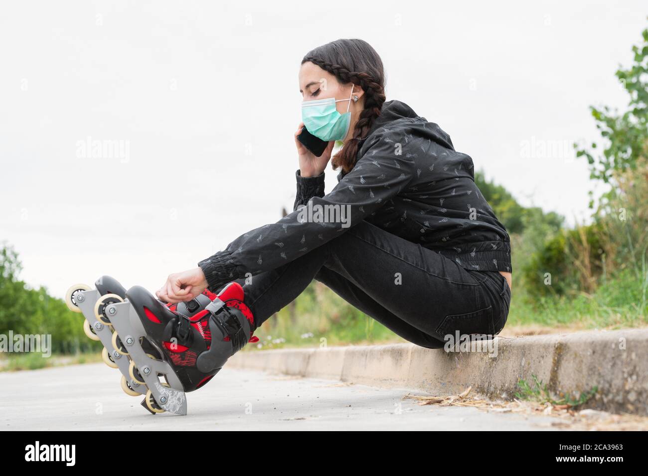 Woman in protective face mask on roller skating pause, sitting on the street and using mobile phone during coronavirus pandemic outbreak. Urban Girl Stock Photo