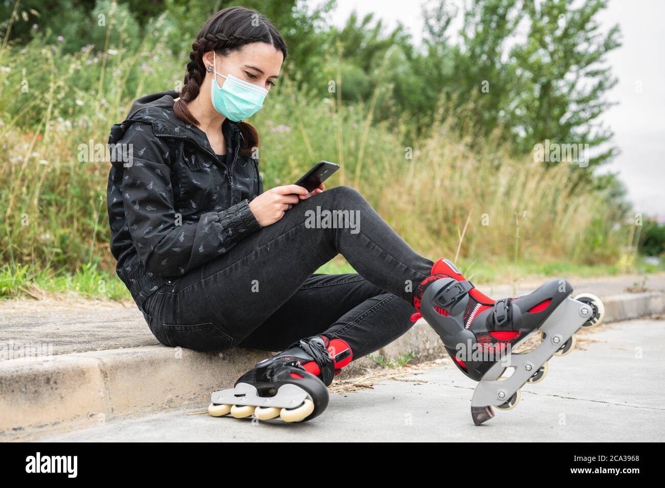 Woman in protective face mask, on roller skating pause, sitting on the street and using mobile phone during coronavirus pandemic outbreak. Urban Girl Stock Photo